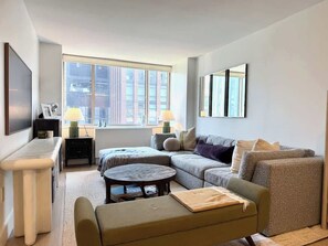 A modern living room with a sofa, armchair, and a small round table. There is a large window providing natural light and a view of buildings outside. - Pied-a-terre in the Sky (NY)