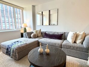 A living room with a sectional sofa, a round coffee table, and a large window. - Nomad Luxury Pied-a-Terre NYC (NY)