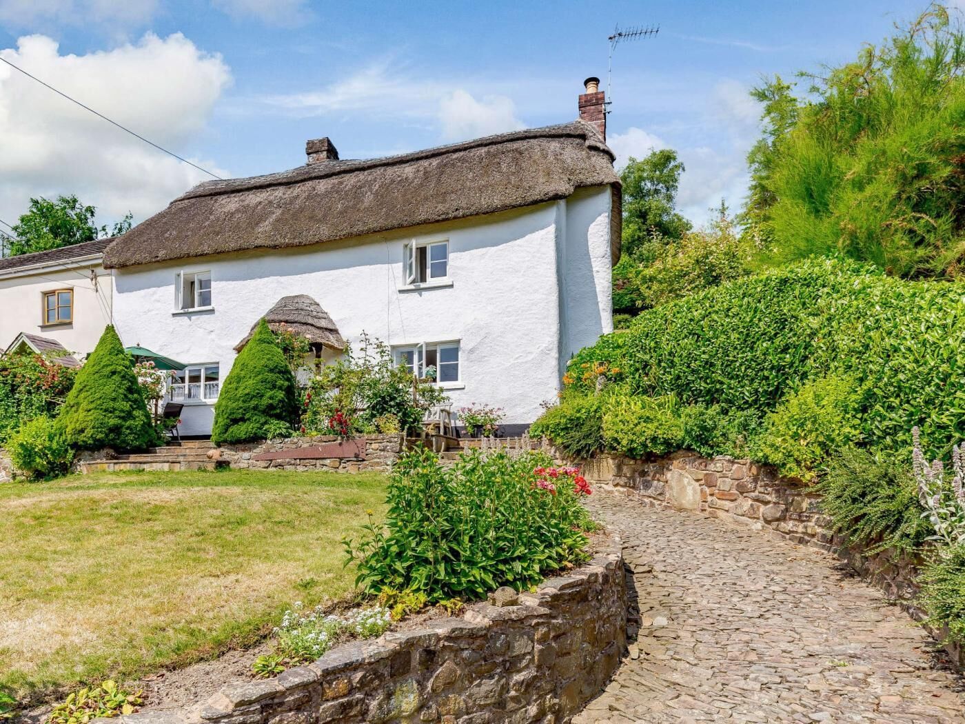 A traditional white house with a thatched roof, surrounded by a stone wall and lush greenery.