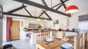 A modern kitchen with a central island, wooden beams, and a dining area with a table set for two. - 4 Bed in Chathill (oc-83469) (Swinhoe)