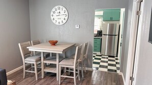 Un coin repas avec une table en bois, quatre chaises, un saladier, une horloge murale et une cuisine visible à travers une porte ouverte.