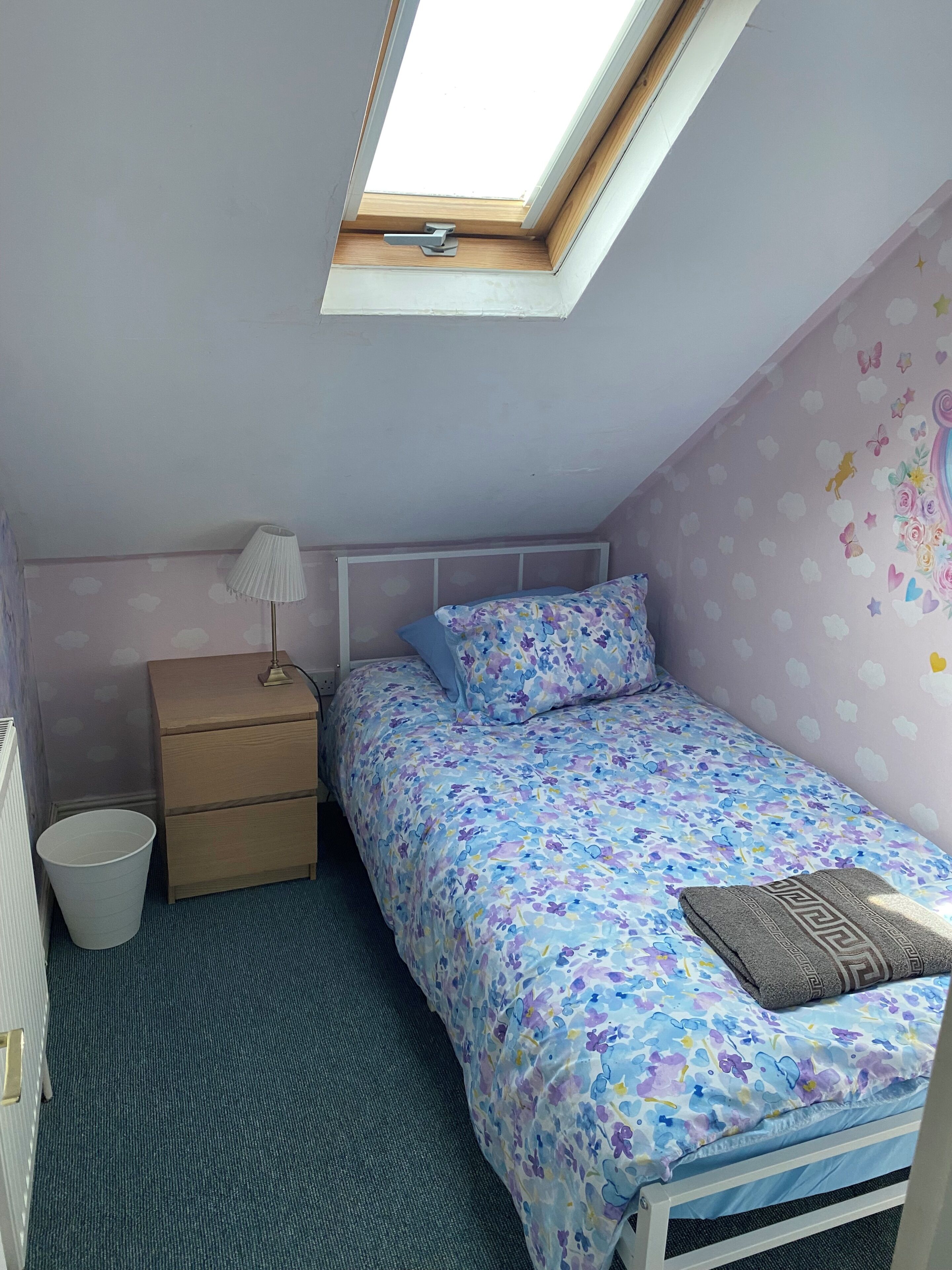 A bedroom with a skylight, a bed with floral bedding, a bedside table with a lamp, and a radiator.