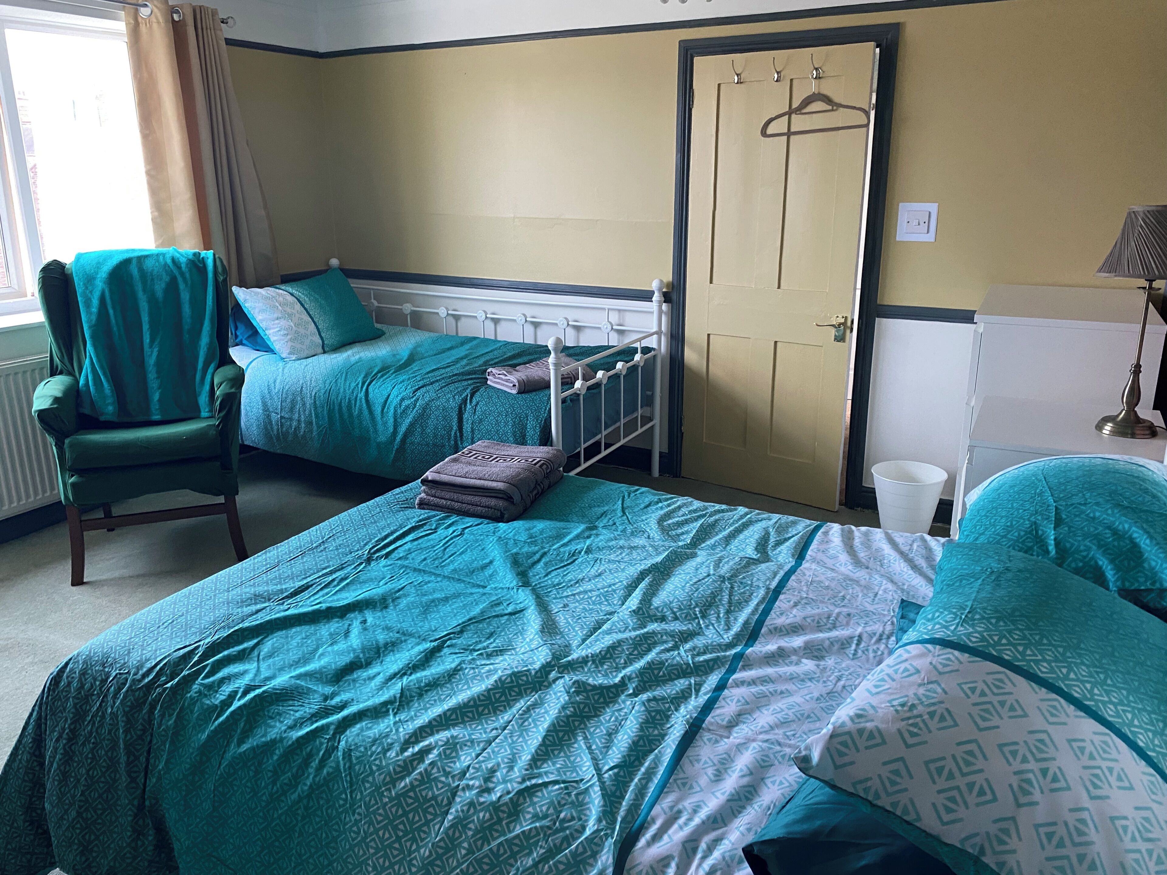 A hotel room with two beds, a chair, a radiator, a door, and a lamp.