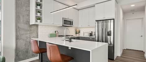 A modern kitchen with white cabinets, a stainless steel refrigerator, a microwave, a sink, and a bar area with two stools.
