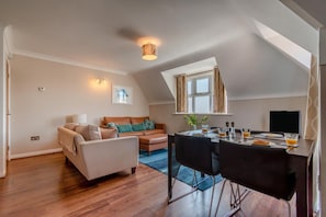 A modern living room with a sofa, a dining table set for breakfast, and a large window with curtains. - 3 Bed in Swanage (oc-83794) (Swanage)
