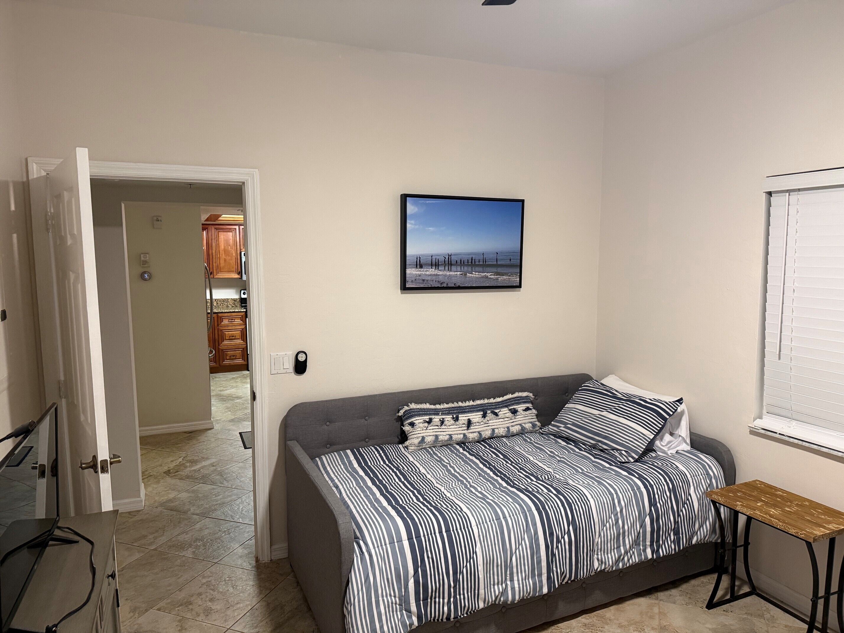 Un dormitorio con un sofá cama gris, ropa de cama a rayas, una pequeña mesa de madera y un cuadro en la pared.