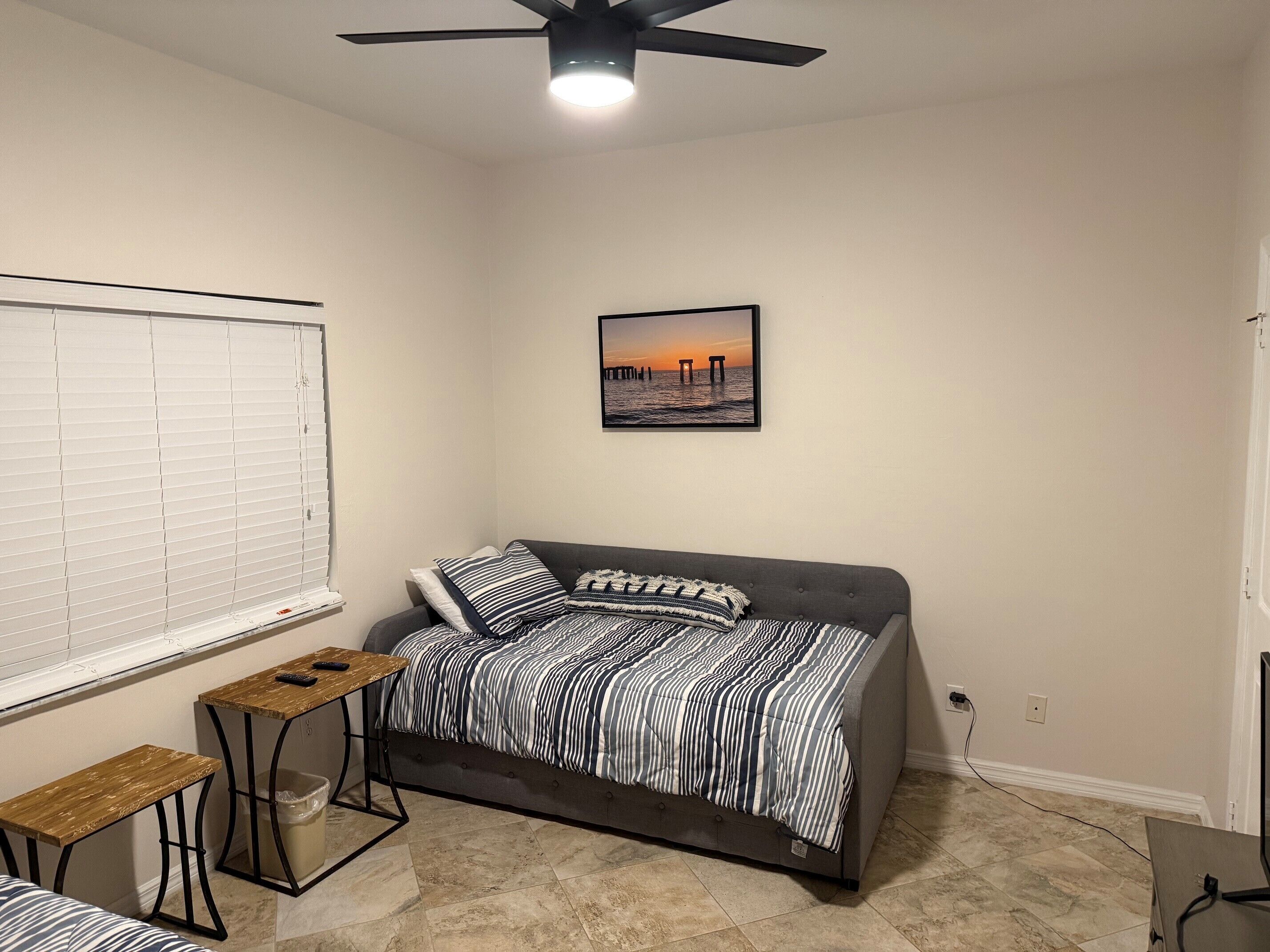 Une chambre à coucher comprenant un lit, une table de chevet, une fenêtre avec des stores, un ventilateur de plafond et un tableau accroché au mur.