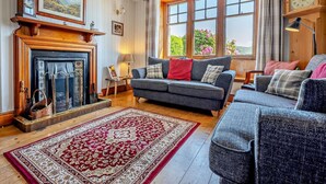 Un salon avec une cheminée, un canapé, un tapis et une grande fenêtre ornée de fleurs.
