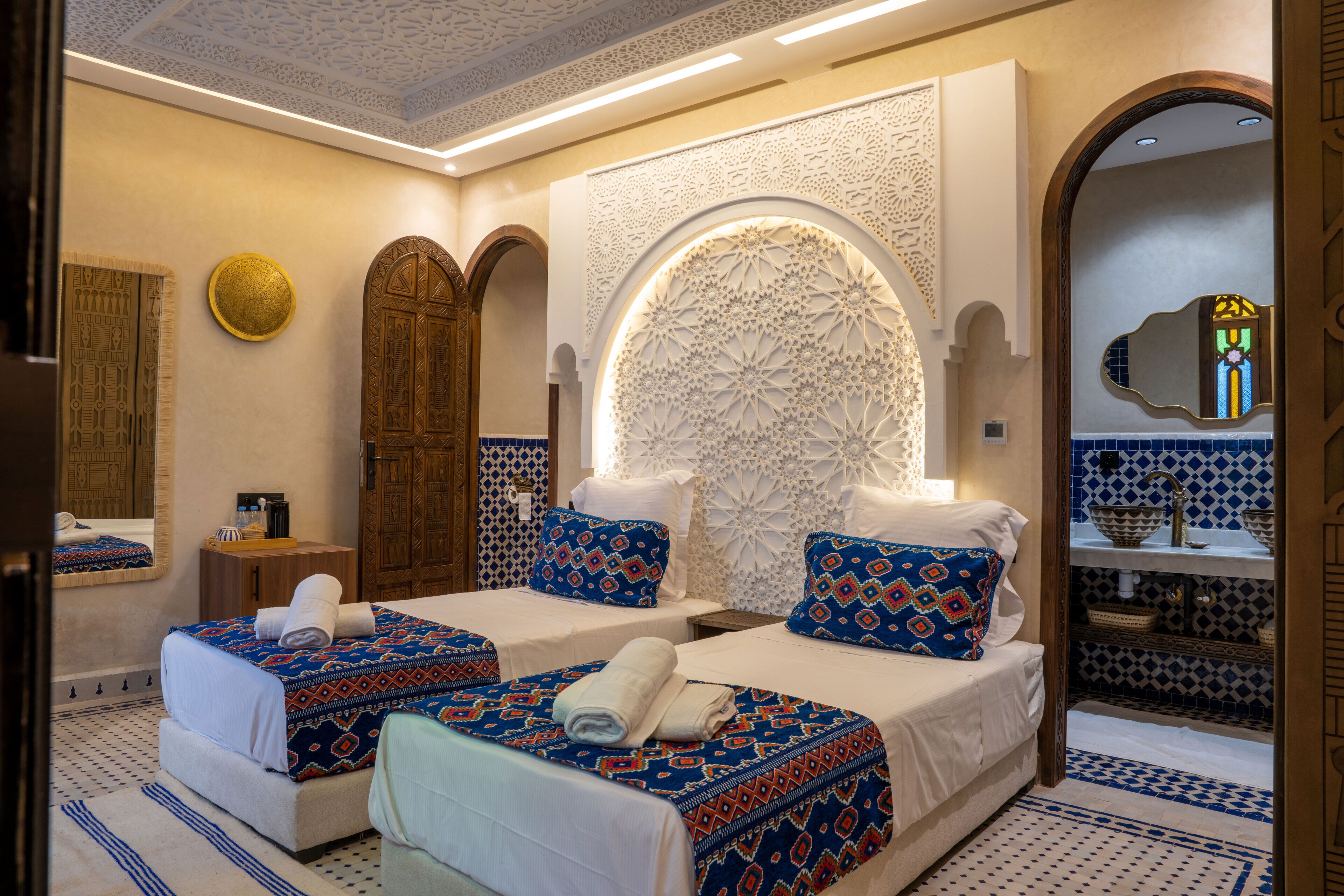 Une chambre d’hôtel avec deux lits, un mur décoratif, un miroir et une salle de bain avec des carreaux à motifs.