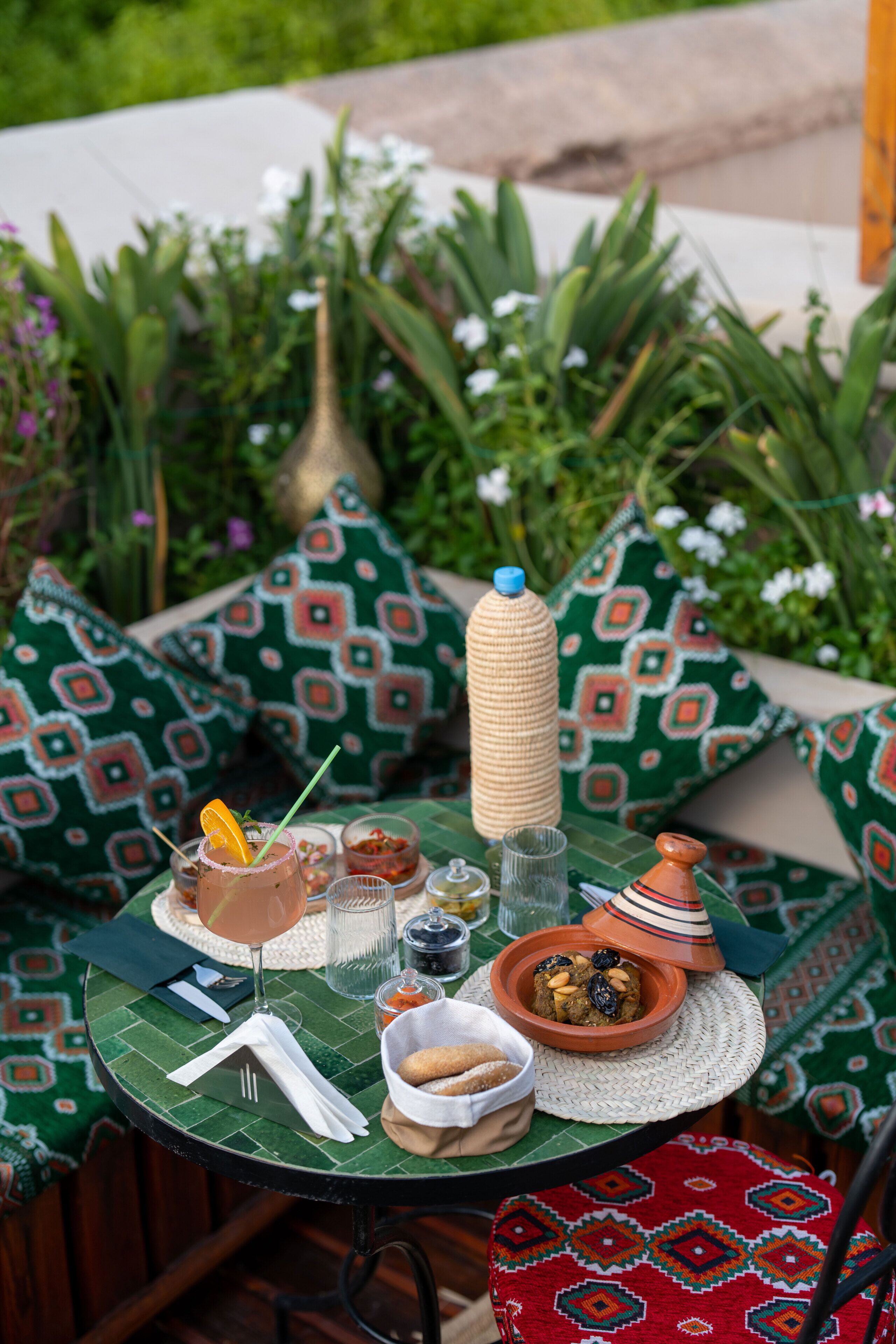 A table set outdoors with a variety of items including a bottle, glasses, a bowl, and a plate, surrounded by patterned cushions.