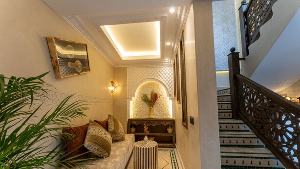 A well-lit hallway with a sofa, decorative wall panels, and a staircase with intricate railing design. - Riad Secret Mahjouba (Marrakech)