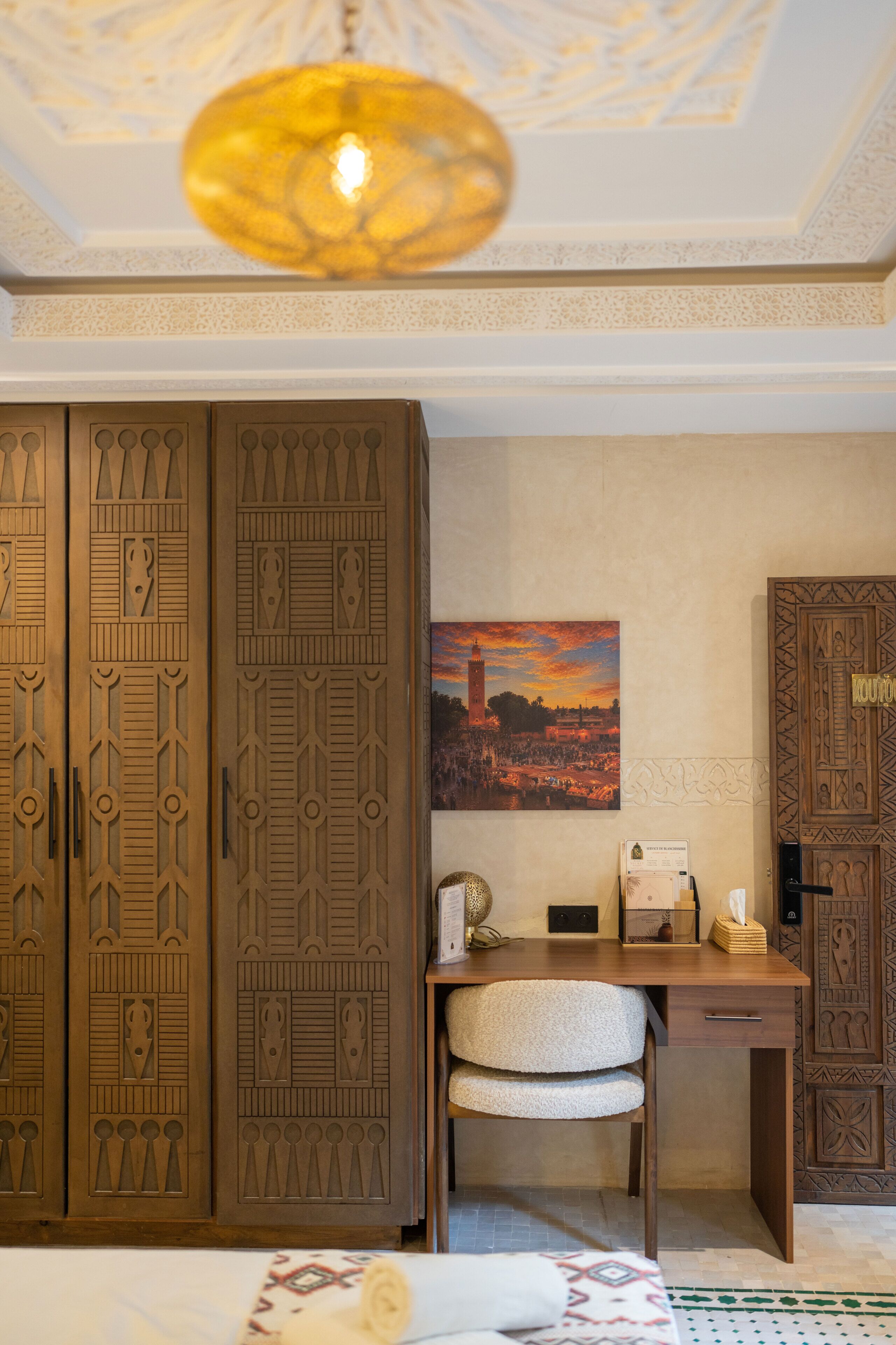 A room with a desk, chair, and a large wooden wardrobe with intricate carvings.