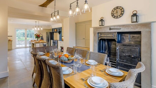 A dining area with a wooden table set for six, surrounded by wicker chairs, a fireplace, and a clock on the wall. - 5 Bed in Harrogate (oc-83892) (Markington)