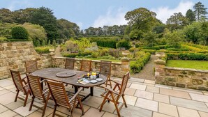 A round wooden table with chairs, a stone wall, and a garden with various plants and a hedge. - 5 Bed in Harrogate (oc-83892) (Markington)