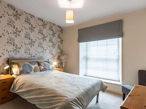 A bedroom with a bed, bedside tables, a lamp, a fan, a window with blinds, and a wooden bench. - 2 Bed in Tenby (oc-83211) (Tenby)