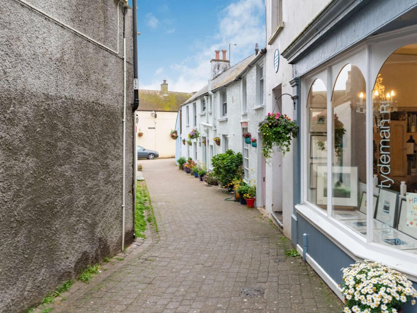A narrow street with white buildings, a paved path, hanging flower baskets, and a visible shop window.