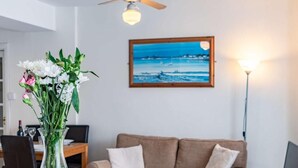 Un salon avec un canapé beige, un tableau représentant un paysage marin, un vase de fleurs et un ventilateur de plafond.