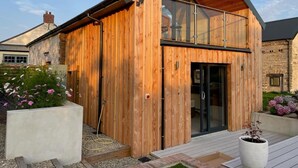 A modern wooden building with a glass entrance, surrounded by a garden with flowers and a small outdoor seating area. - 2 Bed in Cardigan (oc-83919) (Cilgerran)