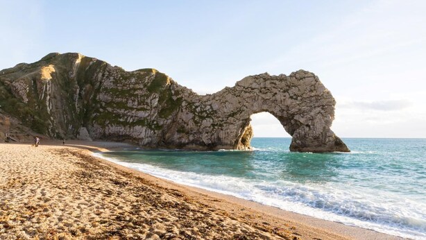 Una playa con un arco natural de roca, cielos despejados y mar tranquilo.