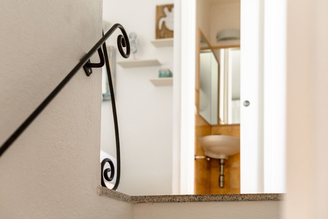Een hoek van een kamer met een witte muur, een zwarre metalen leuning en een glimp van een badkamer met een wastafel en spiegel.