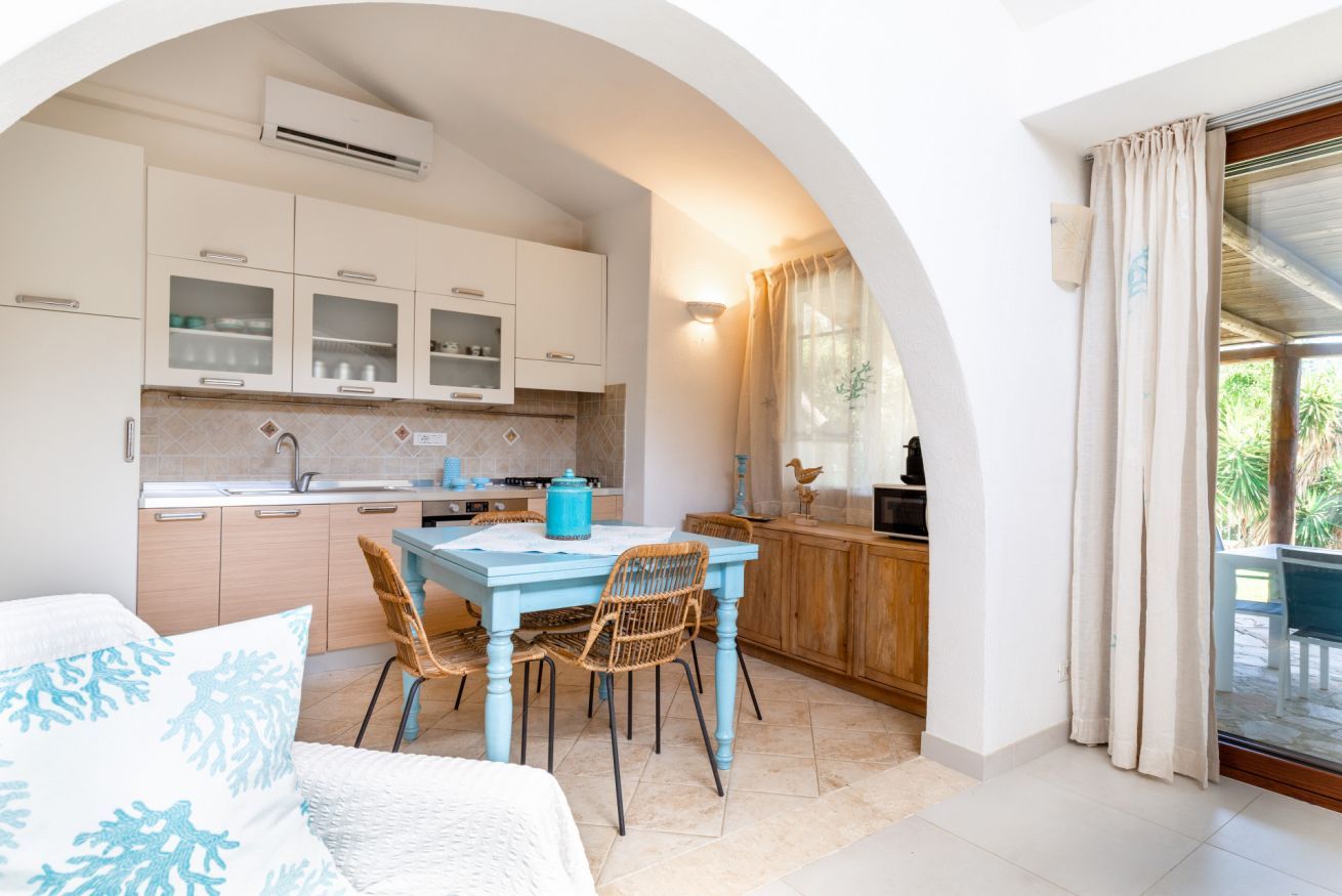 Una cocina compacta con armarios blancos, una mesa azul, sillas de mimbre y un comedor con vistas a través de un arco.