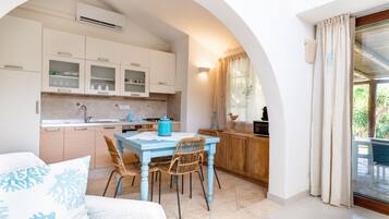 Una cocina compacta con armarios blancos, una mesa azul, sillas de mimbre y un comedor con vistas a través de un arco.