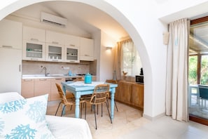 A compact kitchen with white cabinets, a blue table, wicker chairs, and a dining area with a view through an archway. - Sa Barchitta in Villasimius in the bay of Porto Luna (Villasimius)