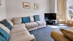A living room with a sofa, a television, a wicker chair, and a window with curtains. - 6 Bed in Keswick (oc-sz389) (Keswick)
