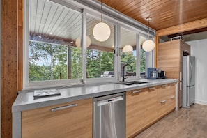 A modern kitchen with a large window, stainless steel appliances, and wooden cabinetry. - Charlevoix Cabin | Hot Tub |Pool Table | 22 Guests (Petite-Rivière-Saint-François)