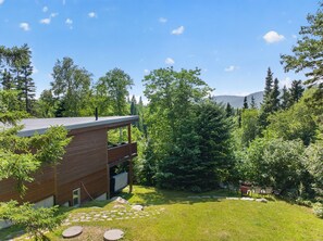 A modern house with a flat roof, a wooden deck, and a stone pathway leading to the entrance. - Charlevoix Cabin | Hot Tub |Pool Table | 22 Guests (Petite-Rivière-Saint-François)