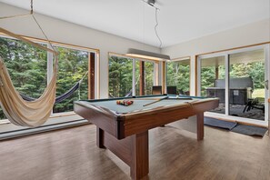 Une table de billard dans une pièce dotée de grandes fenêtres, d’un hamac et offrant une vue sur une forêt.