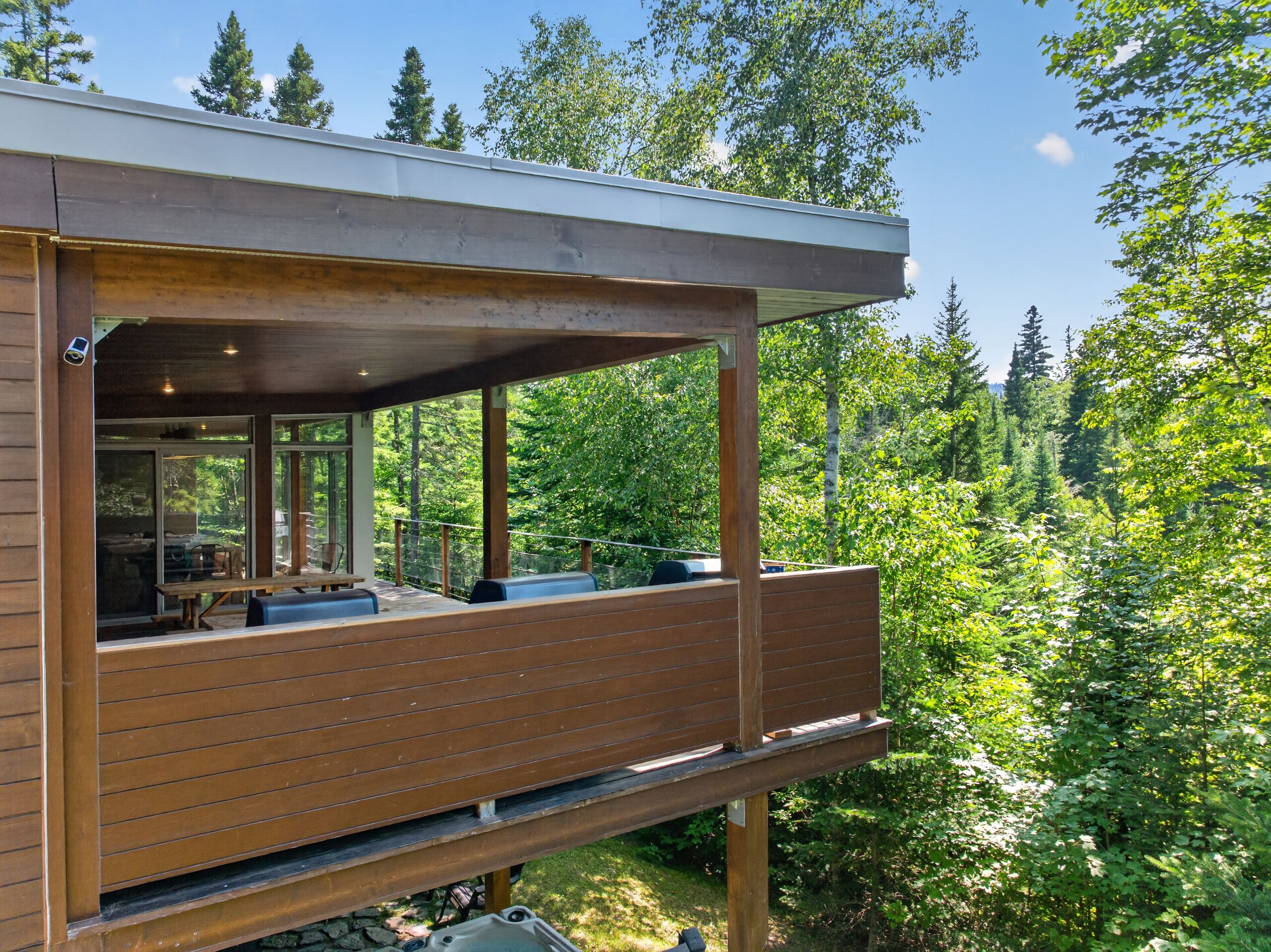 A modern wooden cabin with a covered deck and large windows overlooking a forested area.