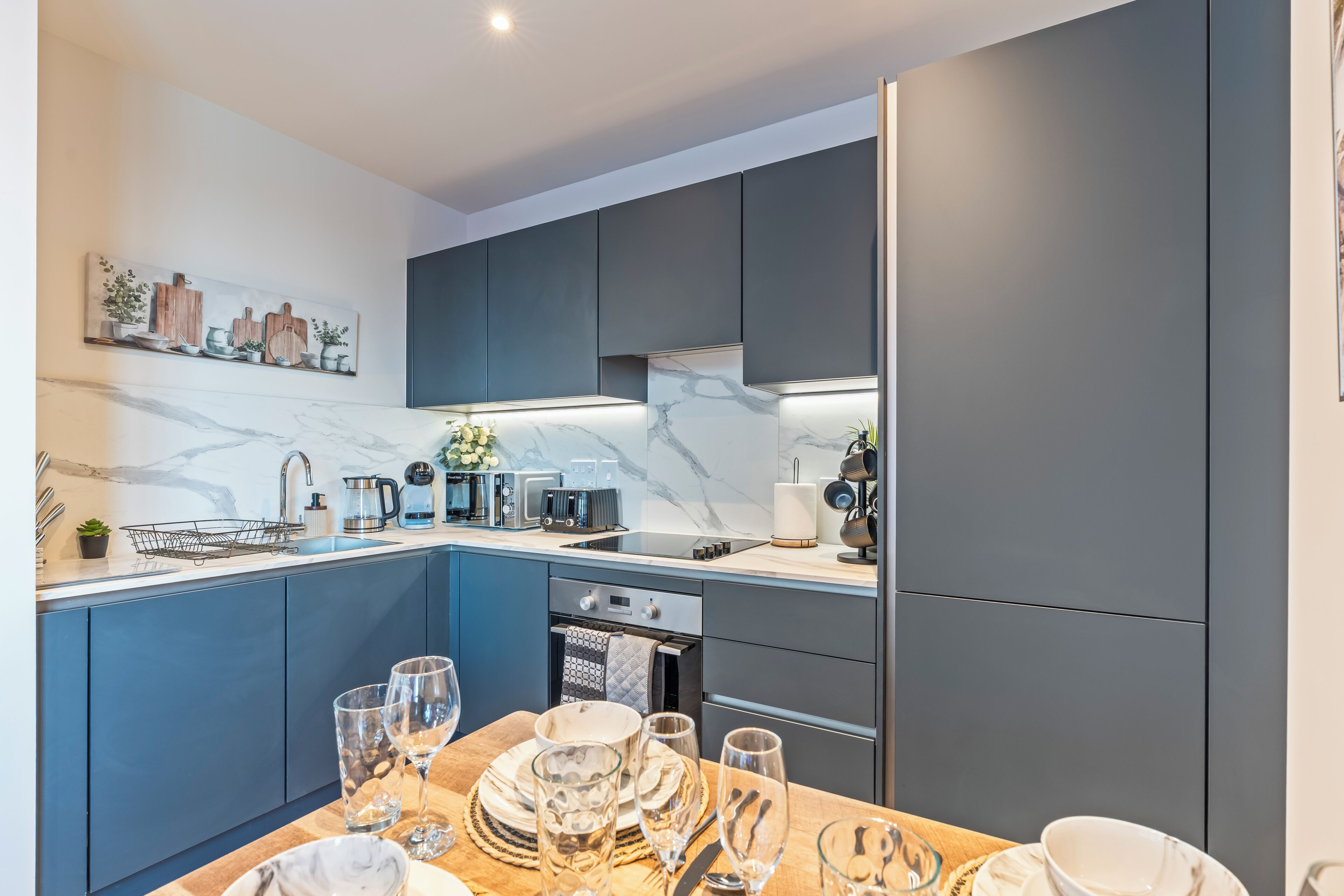 Een moderne keuken met donkerblauwe kastjes, een marmeren aanrechtblad en een eethoek gedekt met borden, glazen en bestek.