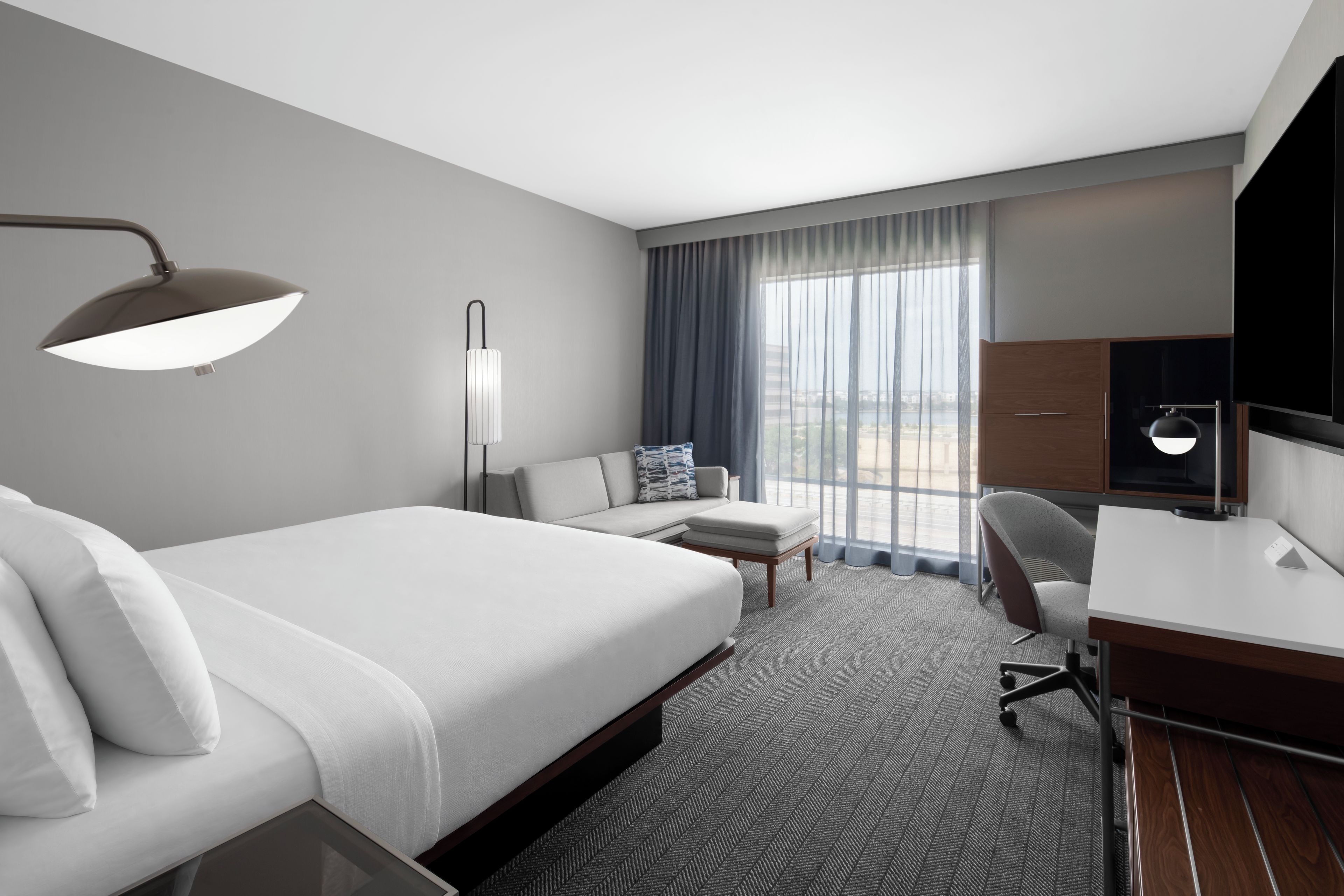 A modern hotel room with a large bed, a desk, a chair, and a view of the city through the window.