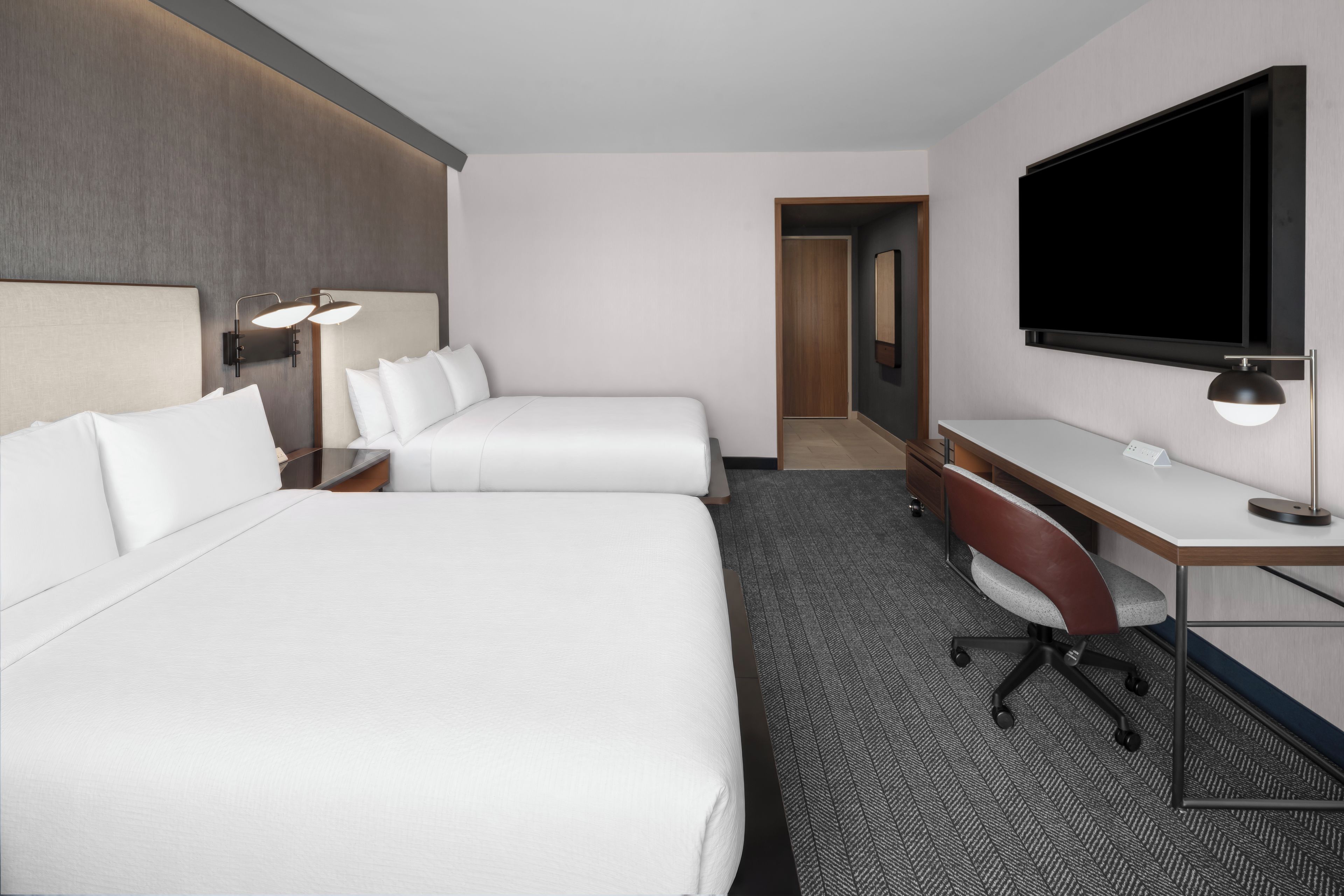 A hotel room with two beds, a desk, a chair, a TV, and a door leading to a bathroom.