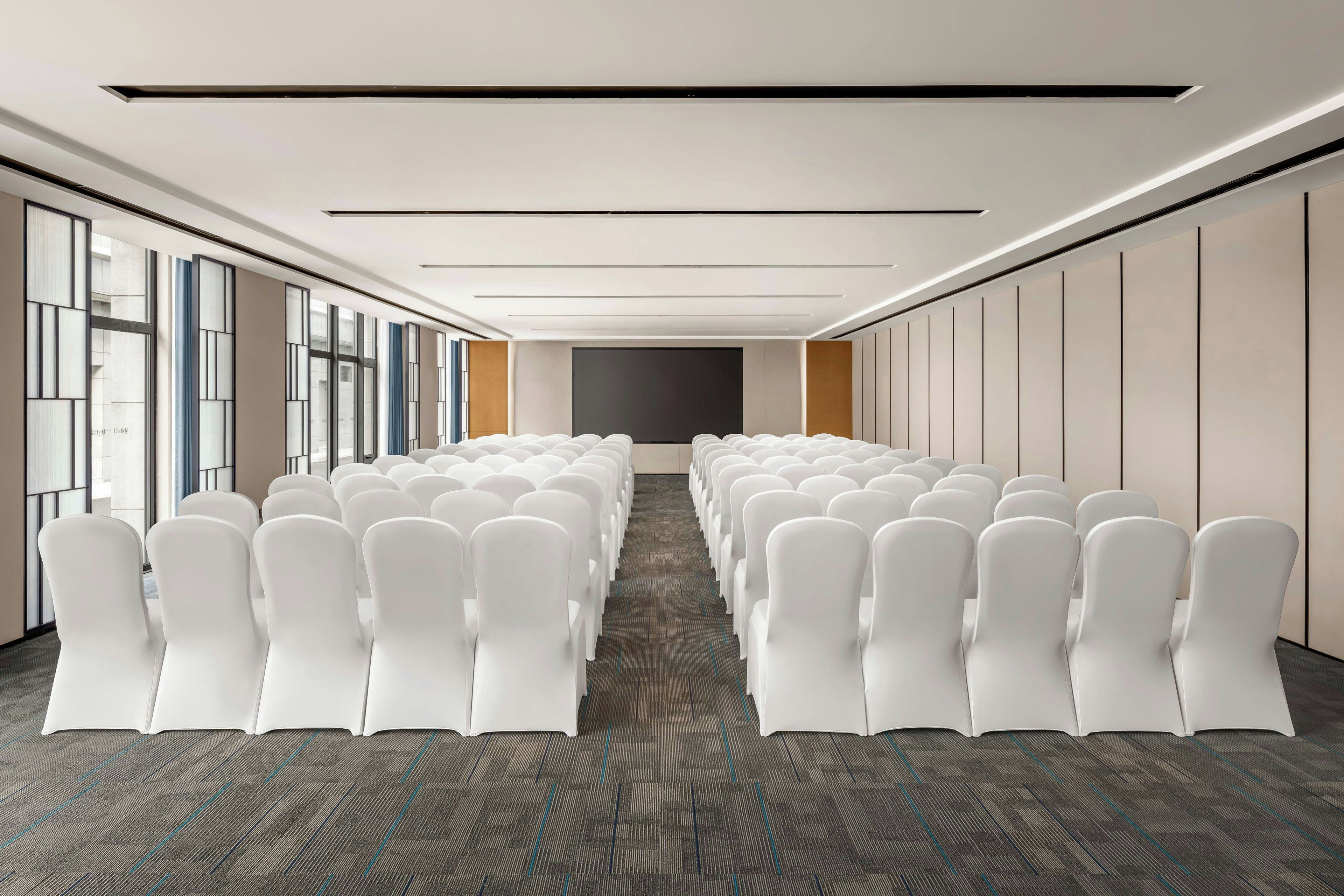 A conference room with rows of white chairs, a carpeted floor, and a large window on one side.