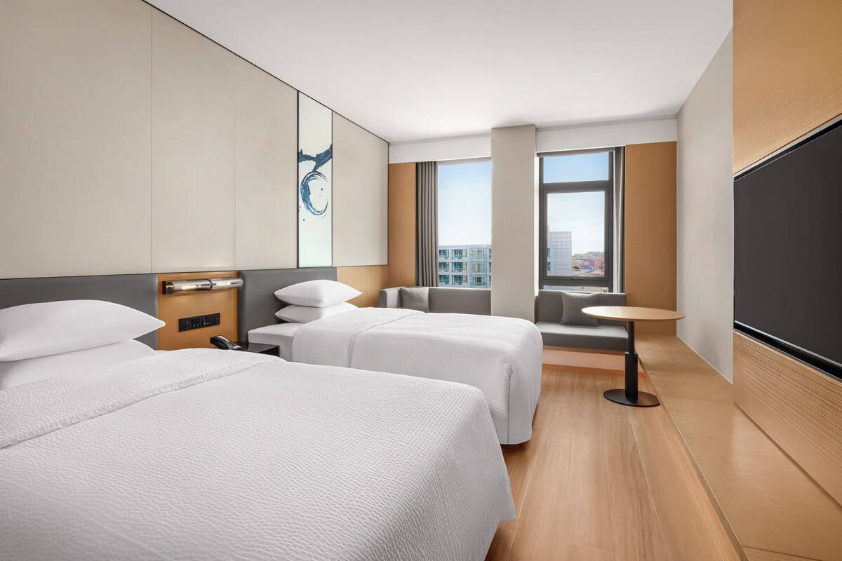 A hotel room with two beds, a small table, a sofa, and a large window offering a city view.