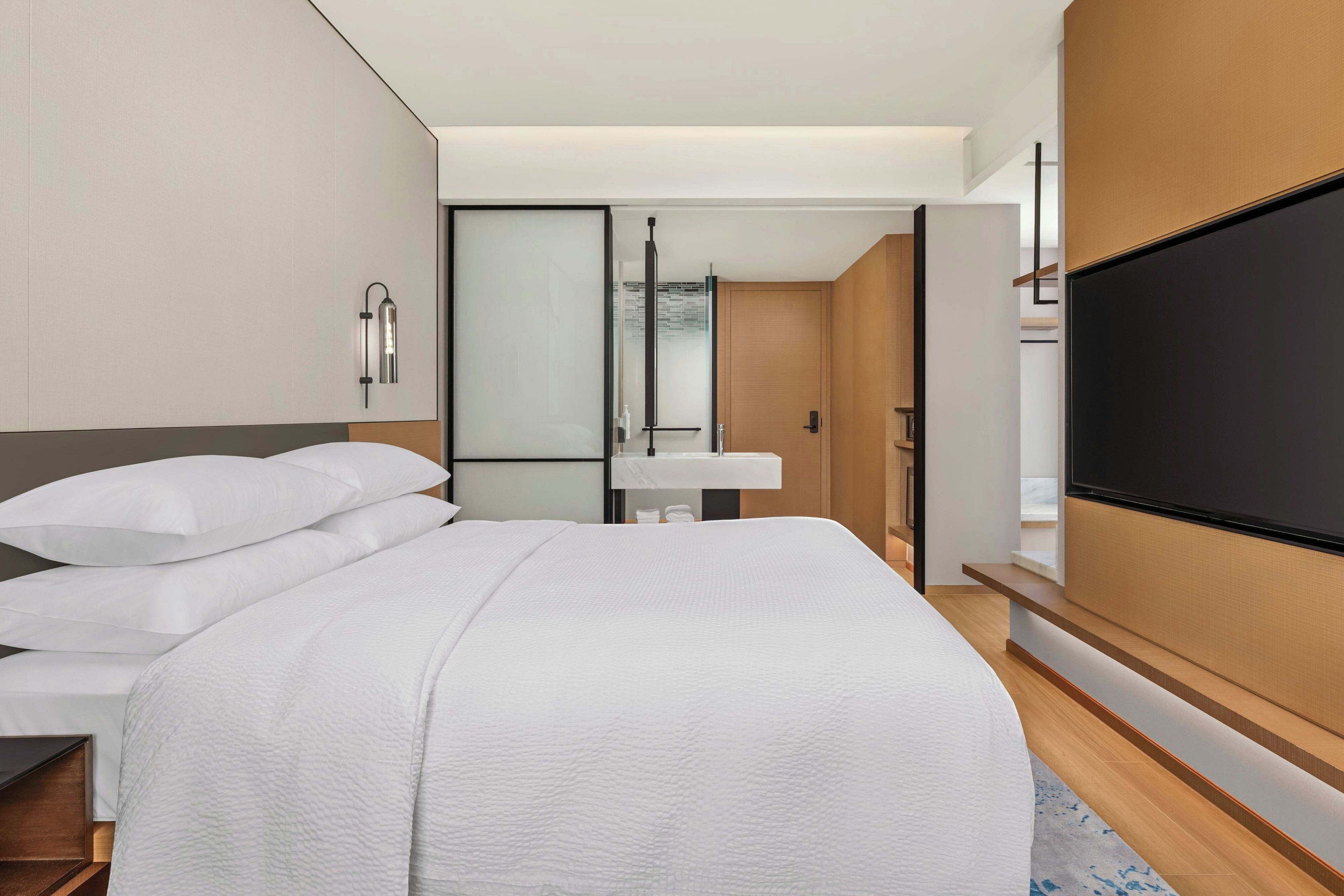 A modern hotel room with a large bed, a flat-screen TV, a bathroom with a mirror, and a sliding door leading to another room.