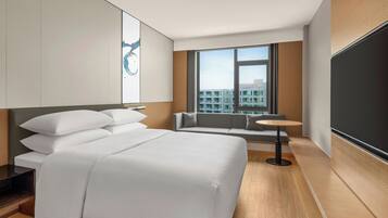 Una habitación de hotel moderna con una cama grande, un sofá pequeño, una mesa redonda y un amplio ventanal con vistas a la ciudad.