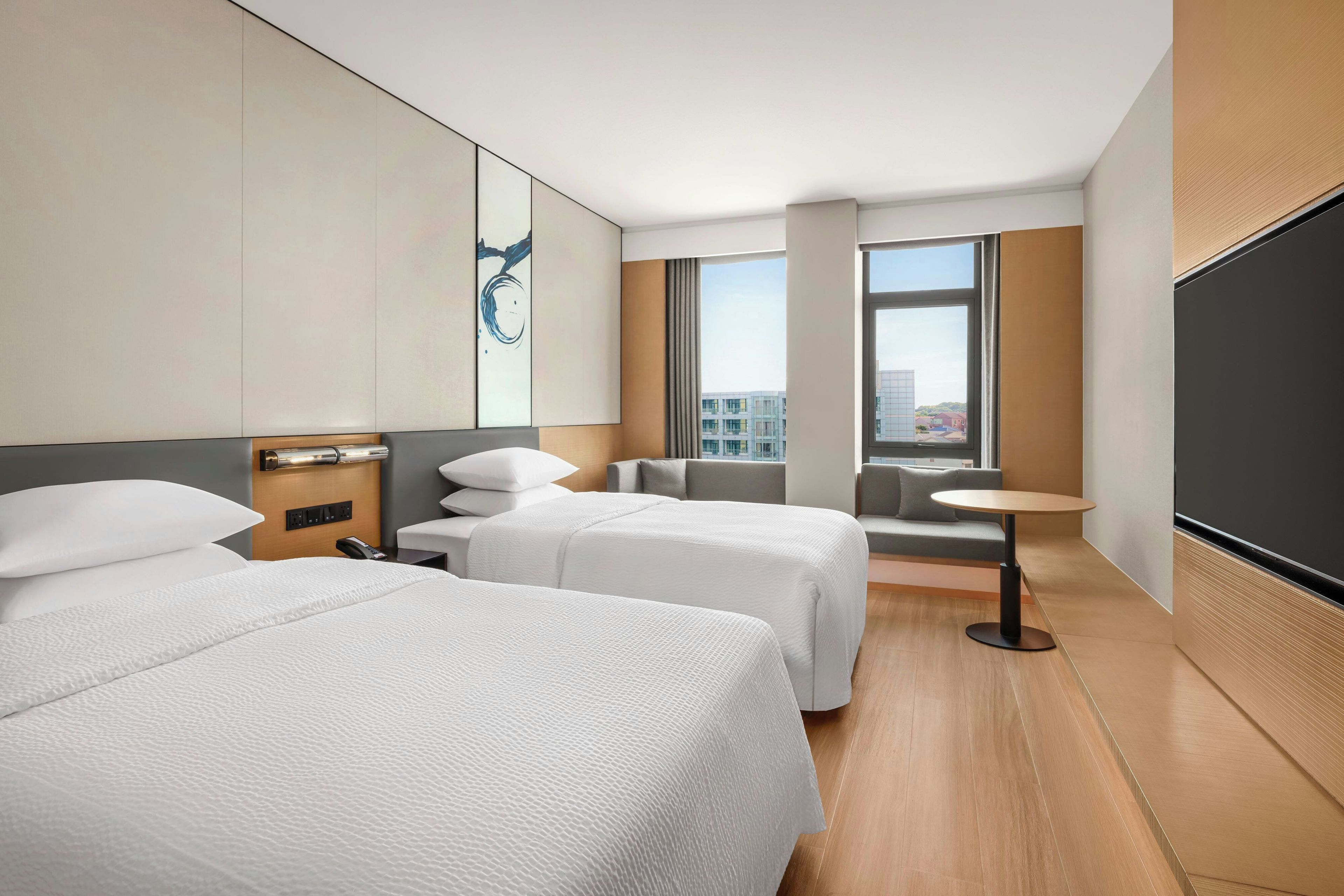 A hotel room with two beds, a small table, a sofa, and a large window offering a city view.