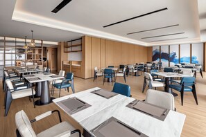 Un comedor moderno con varias mesas y sillas, una zona de cocina con armarios, y un ventanal con vistas.
