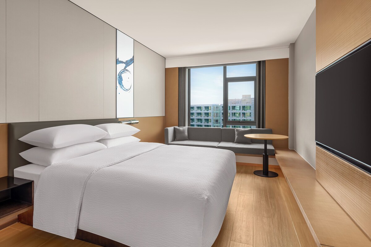 A modern hotel room with a large bed, a sofa, a small table, and a large window offering a city view.