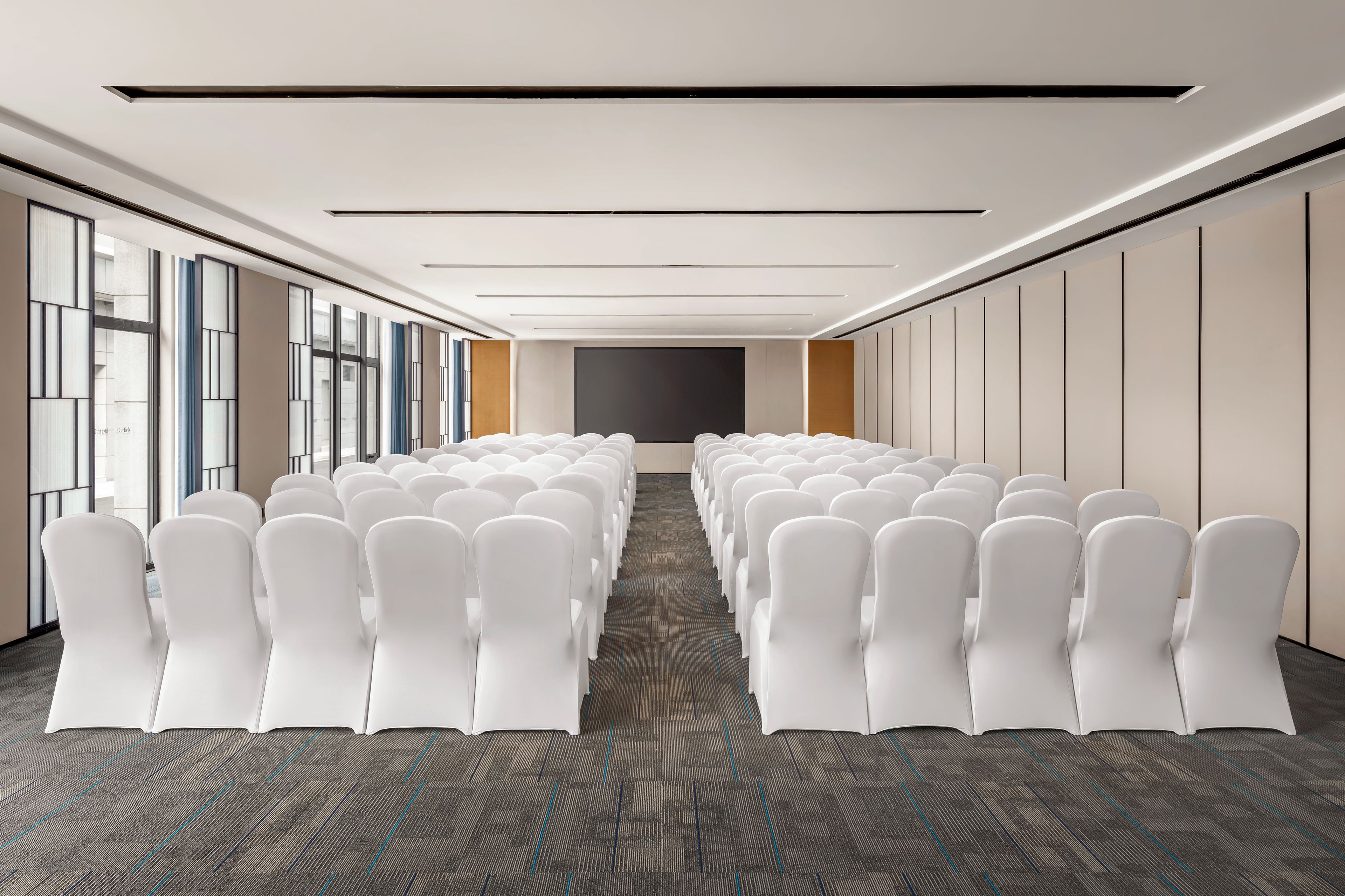A conference room with rows of white chairs, a carpeted floor, and a large window on one side.