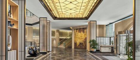 A spacious lobby with a geometric patterned floor, a central staircase, and a large illuminated ceiling.
