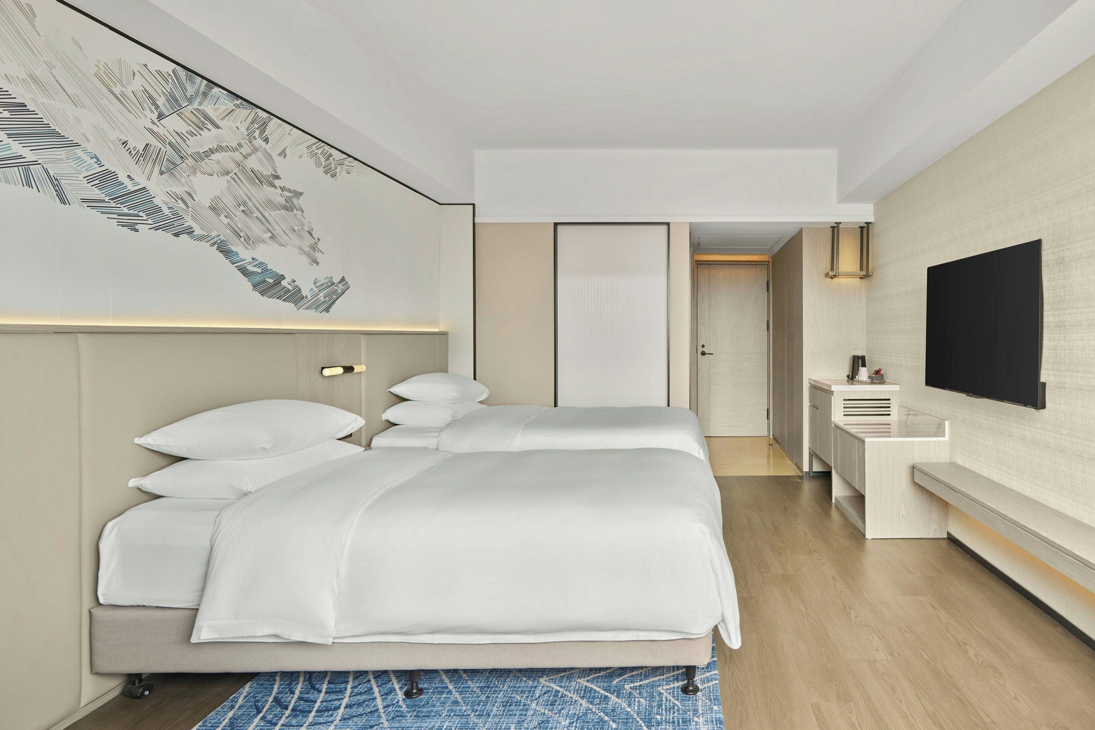 Quarto de hotel com uma cama grande, mesas de cabeceira, escrivaninha e televisão na parede.
