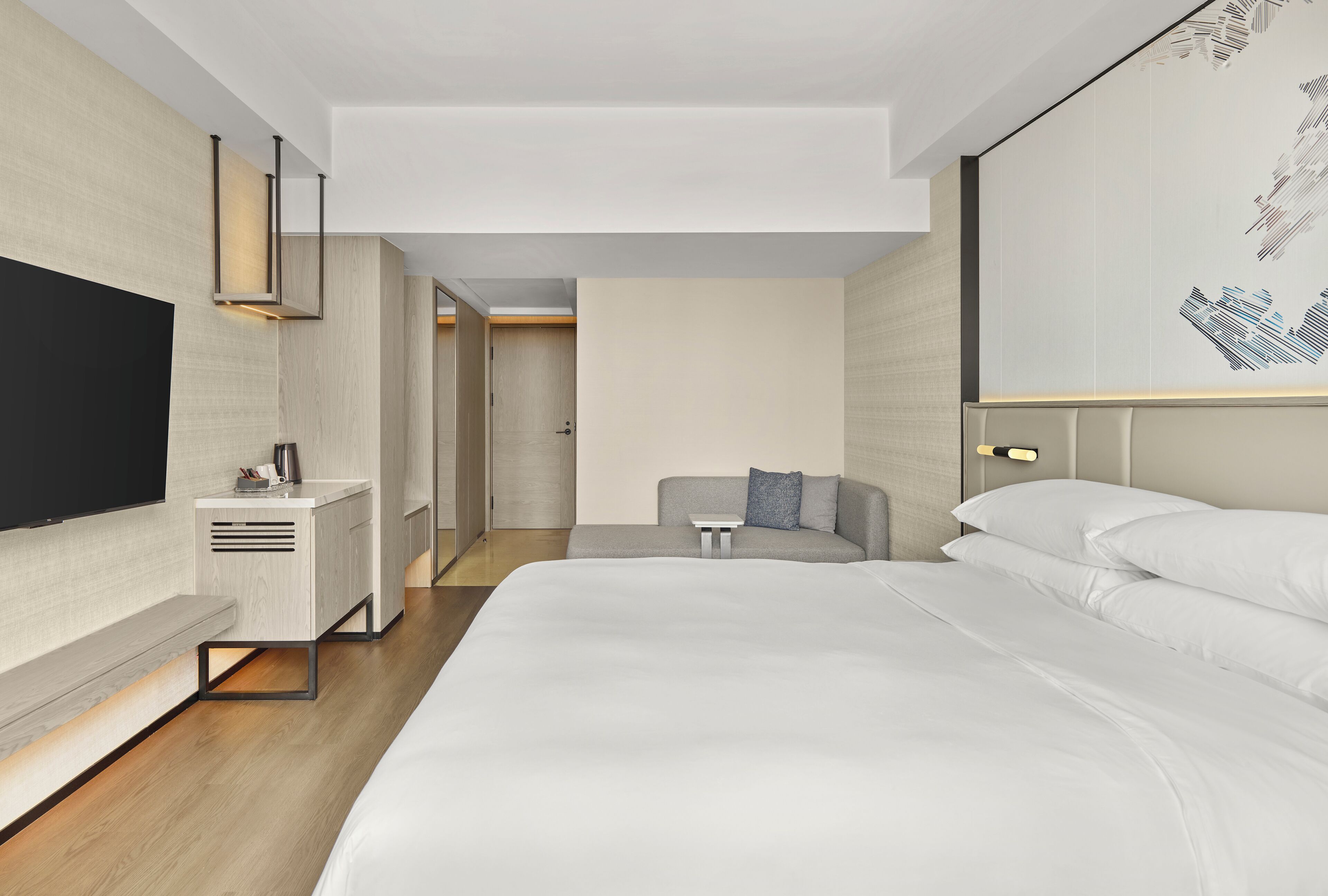 Una habitación de hotel moderna con una cama grande, un televisor de pantalla plana, una mesita de noche y una zona de estar con un sofá y una pequeña mesa.