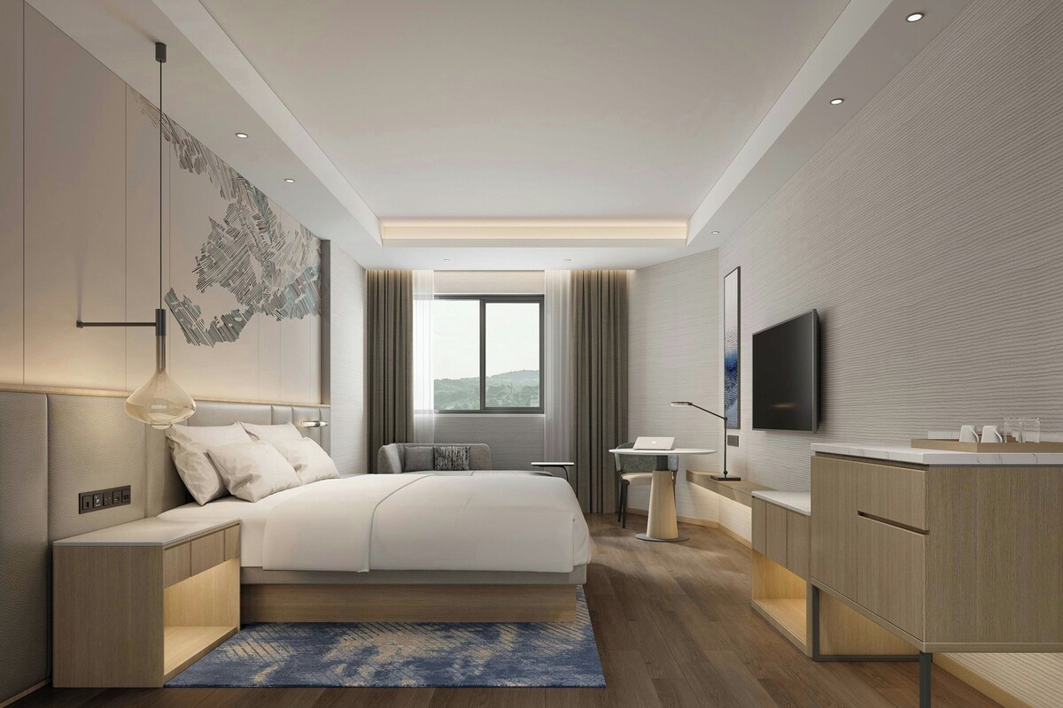 Habitación de hotel moderna con una cama grande, mesitas de noche, un escritorio, un televisor y vistas al exterior.