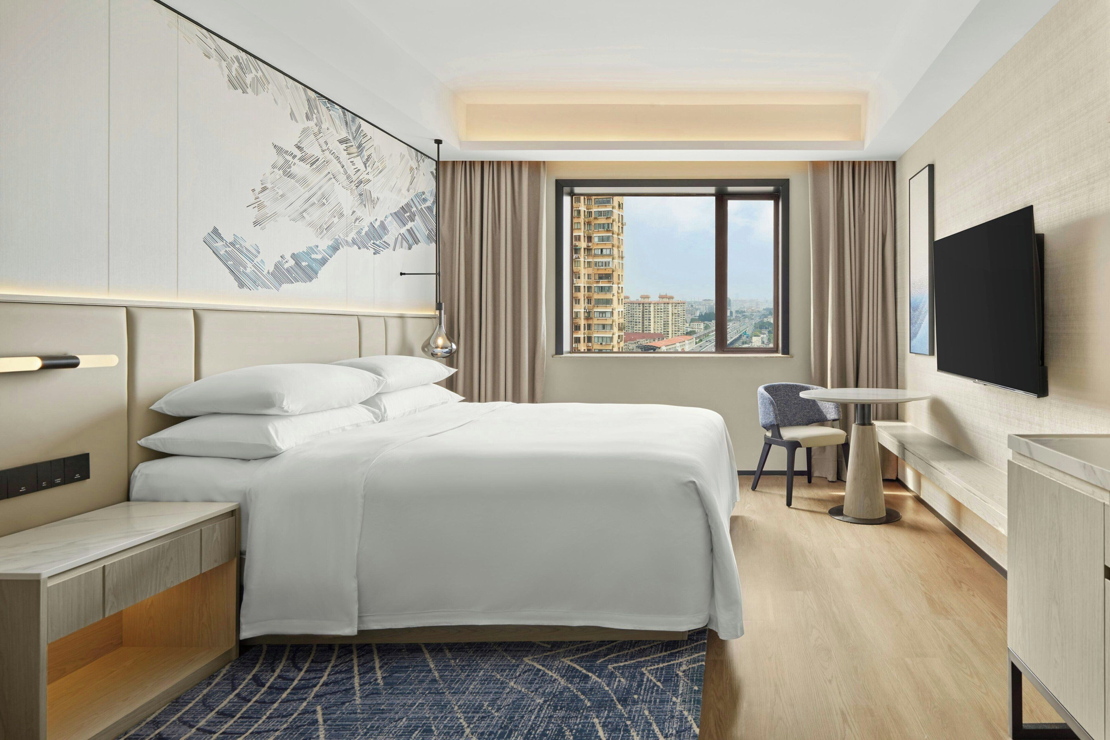 Habitación de hotel con cama, mesita de noche, escritorio, silla y un gran ventanal con vistas a edificios.