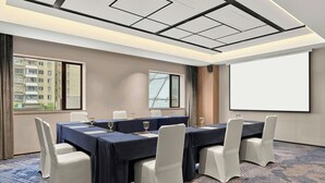 A conference room with a large table, white chairs, a projector screen, and a view of a building through the window.