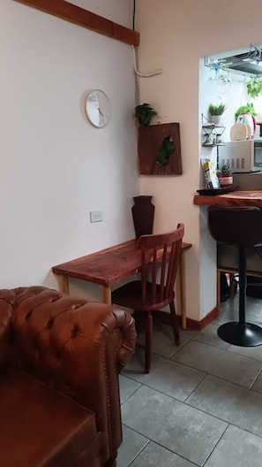 Ein Essbereich mit Holztisch und -stühlen, einer Wanduhr, einem Topf mit Pflanze und einer Küchenzeile mit Mikrowelle und Kaffeemaschine.